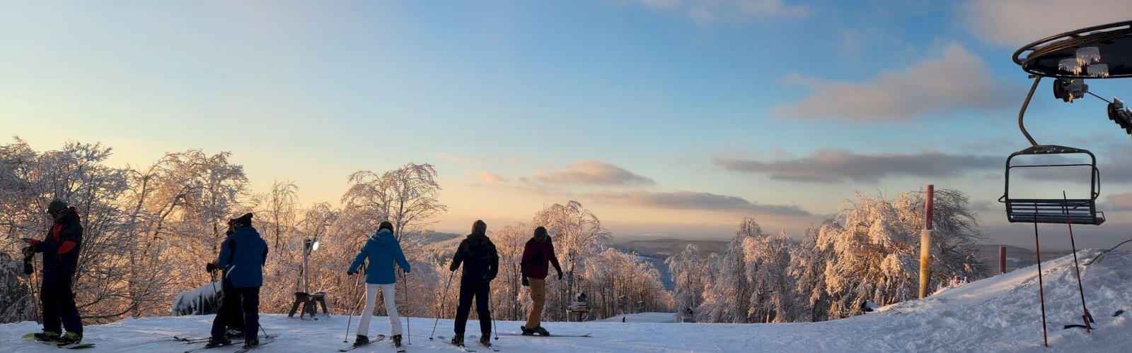 A snowy mountain scene with six skiers standing on a slope at sunset; a ski lift is on the right, and frosted trees glisten in the background.