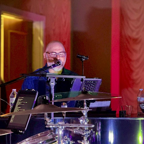 A musician is playing the piano and singing into a microphone, with a music stand and drums nearby.