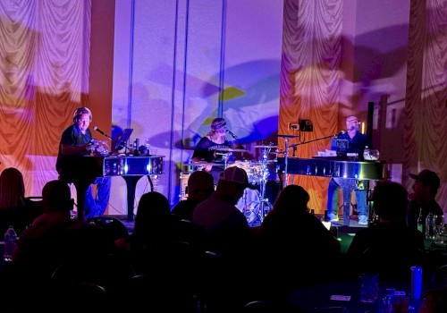 A band performs in a dimly lit room with an audience, featuring two keyboard players and a drummer on stage.