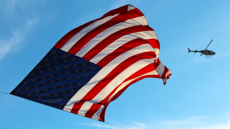 An American flag waving with a helicopter flying in the sky in the background.
