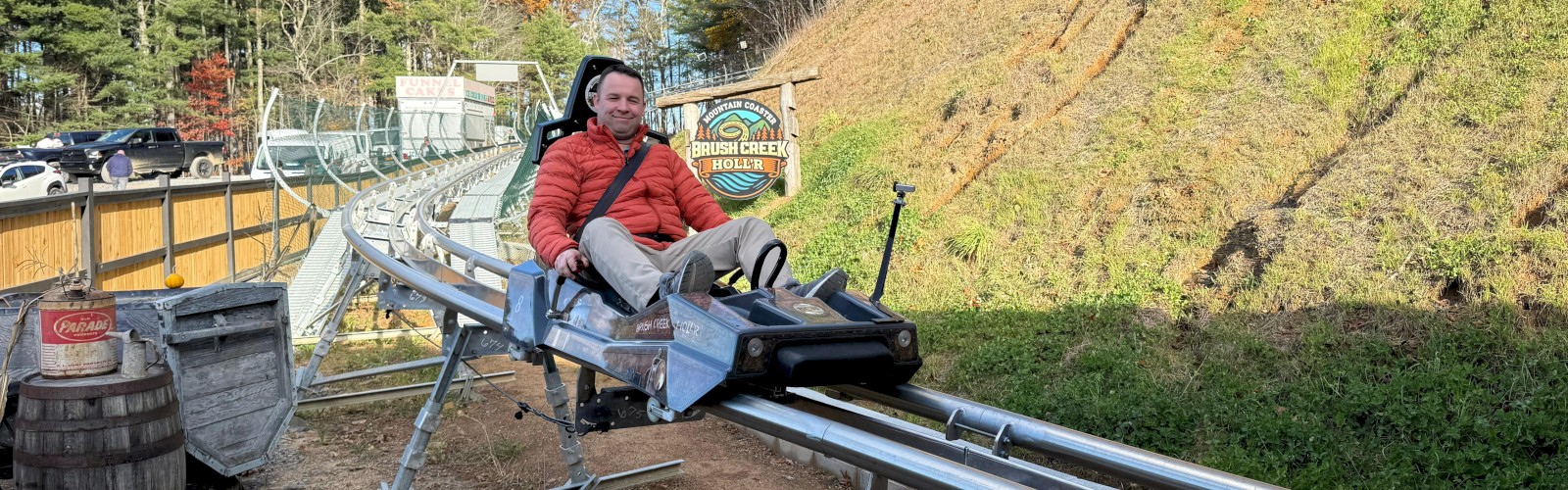 A person is sitting on a mountain coaster ride at Brush Creek Hollow on a clear, sunny day with trees and parked cars in the background.