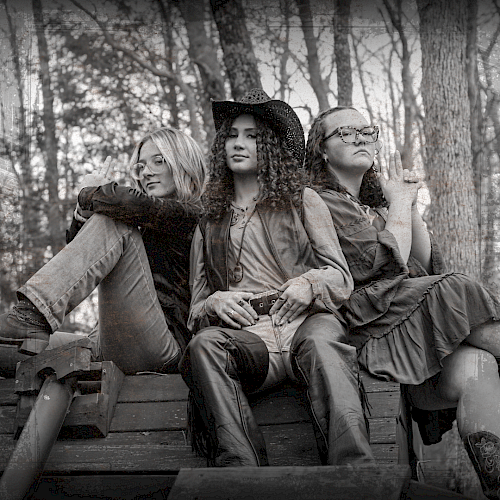 Three women sit on a bench in a forest, striking confident poses; the image is in black and white, highlighting their stylish outfits.
