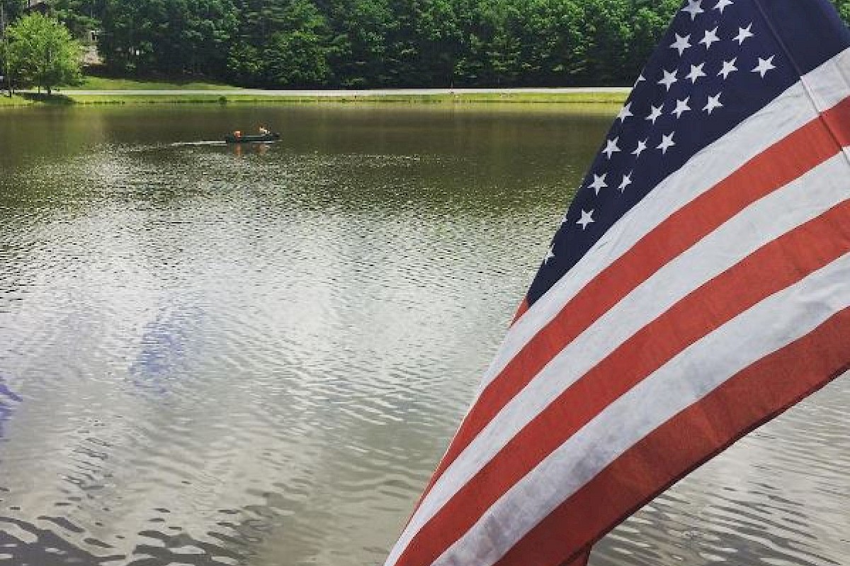 An American flag near a lake with trees in the background and people kayaking in the water.