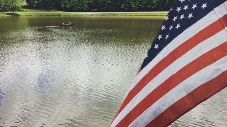 An American flag near a lake with trees in the background and people kayaking in the water.