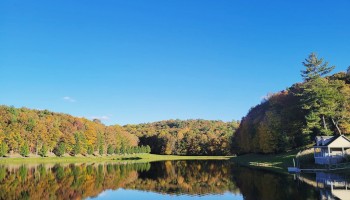 A peaceful lakeside scene with colorful autumn trees, a clear blue sky, and a house by the water reflecting in the calm lake.