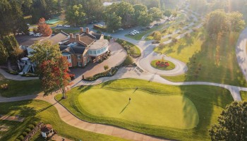 An aerial view of a golf course with a person walking on the green, surrounded by trees, a clubhouse, parking lot, and walking paths.