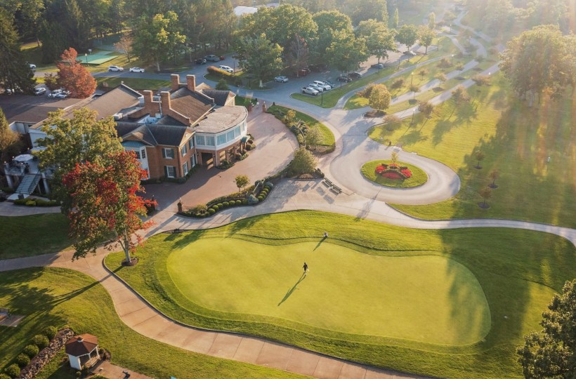 An aerial view of a golf course with a person walking on the green, surrounded by trees, a clubhouse, parking lot, and walking paths.