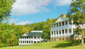 Two large, white, multi-story buildings with green roofs are set on a grassy hill, surrounded by trees and a partly cloudy sky.