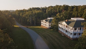 Two residential buildings sit among trees with leaf colors suggesting early fall, along a winding road in a peaceful, wooded area.