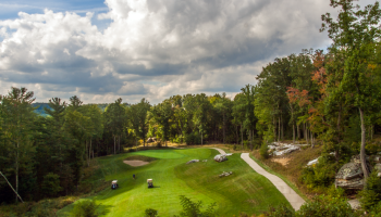This is a scenic golf course surrounded by trees, with carts on the fairway and a cloudy sky overhead.