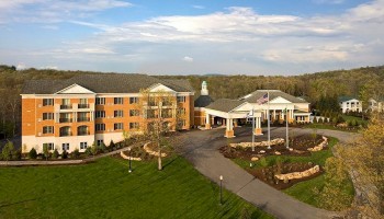 A large, modern hotel or retirement community surrounded by lush greenery, with a driveway, flags, and well-maintained landscaping.