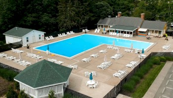 Aerial view of a swimming pool area with lounge chairs, umbrellas, a small building, and surrounding greenery.