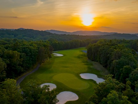A scenic golf course at sunset surrounded by lush trees and rolling hills with sand traps and a winding path.