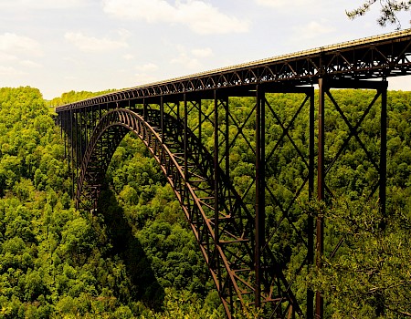 A tall, yellow bridge with an arch spans over a lush green forested valley under a partly cloudy sky.