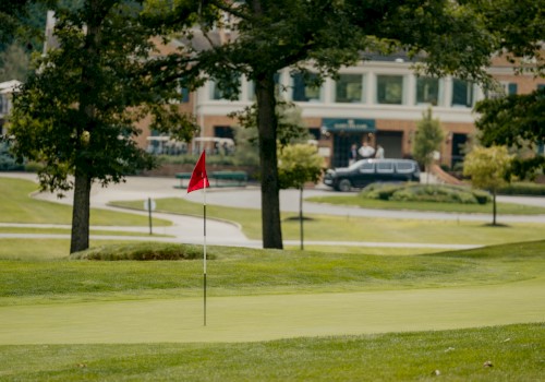 A golf course with a green, flag, trees, and a building in the background on a sunny day.