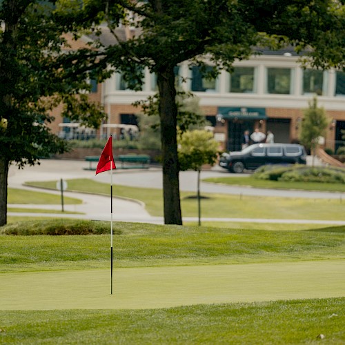 A golf course with a green, flag, trees, and a building in the background on a sunny day.
