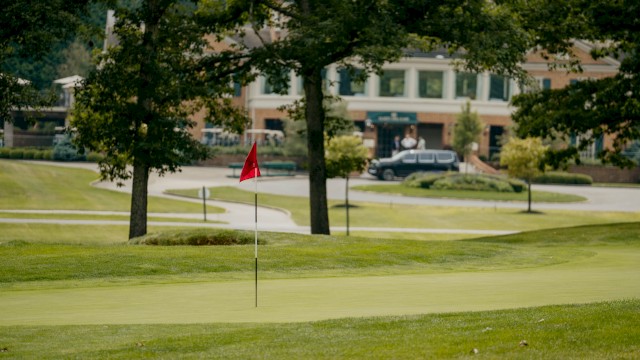 A golf course with a green, flag, trees, and a building in the background on a sunny day.