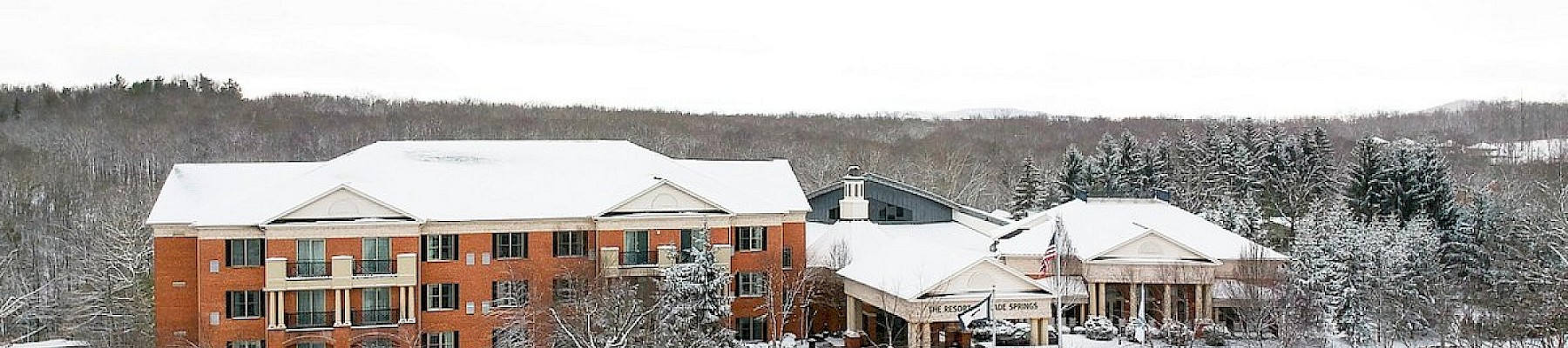 A snowy resort building with a parking lot, trees, and a winding driveway, surrounded by snow-covered landscape and distant forest.