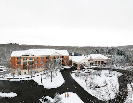 A snowy resort building with a parking lot, trees, and a winding driveway, surrounded by snow-covered landscape and distant forest.