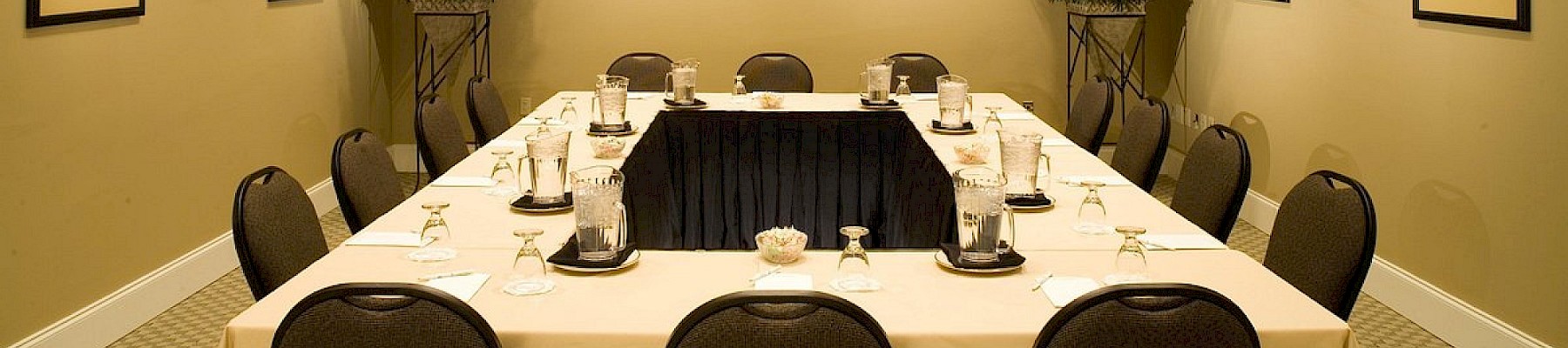 A formal meeting room with a U-shaped table set with glasses, water pitchers, and notepads, decorated with framed black-and-white portraits on beige walls.