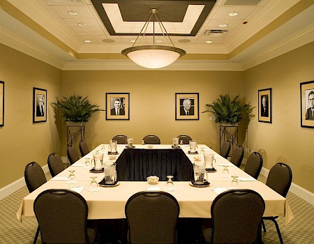 A formal meeting room with a U-shaped table set with glasses, water pitchers, and notepads, decorated with framed black-and-white portraits on beige walls.