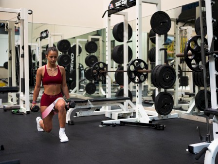 A woman is working out with dumbbells in a gym, kneeling on one knee while holding weights near her leg amidst gym equipment.