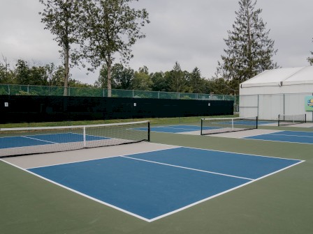 This image shows an empty pickleball court complex with three courts, a white tent structure, and trees, under a cloudy sky.