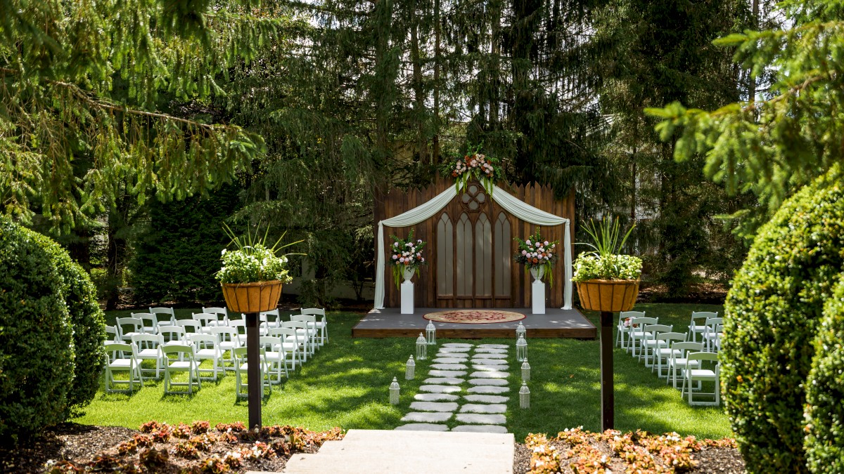 A small outdoor wedding setup with chairs, floral arrangements, and a decorated wooden backdrop in a lush garden.