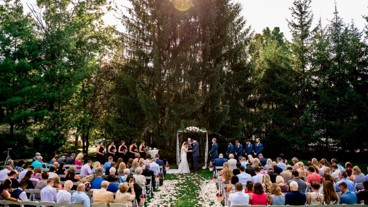 A wedding ceremony takes place outdoors under tall trees with guests seated, officiant, bride and groom, and bridal party in view.