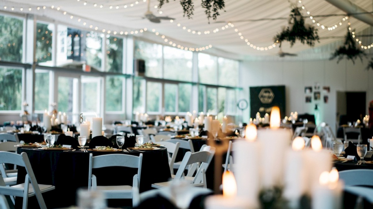 A decorated banquet hall with candlelit tables, string lights, hanging greenery, and large windows letting in natural light.