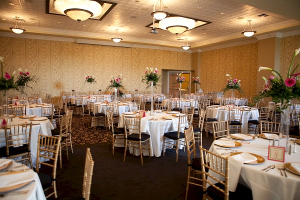 This is an elegantly decorated banquet hall with round tables, floral centerpieces, and gold chairs arranged for an event.