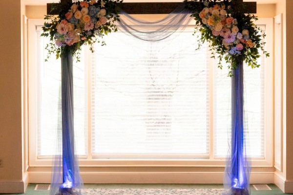 A decorated wedding arch with flowers and fabric stands in front of a large window with blinds.