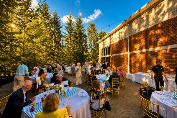 People gather at a beautifully decorated outdoor event with tables covered in white and blue striped tablecloths, enjoying a sunny evening.