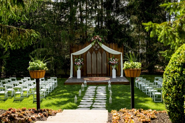 An outdoor wedding setup with chairs facing a decorated wooden backdrop, floral arrangements, and a small aisle leading to the stage.