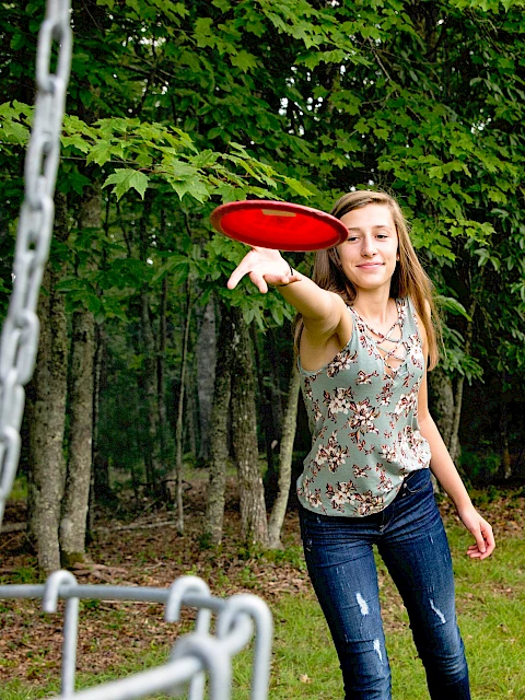 A girl is playing disc golf outdoors in a forested area, reaching to throw a red disc towards a metal chain basket.