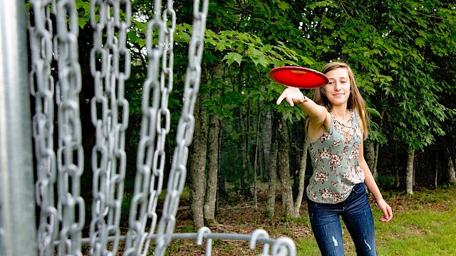 A girl is playing disc golf outdoors in a forested area, reaching to throw a red disc towards a metal chain basket.