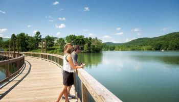 A couple stands on a wooden boardwalk overlooking a calm lake, surrounded by lush green hills under a clear blue sky.