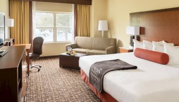 A hotel room with a bed, sofa, desk chair, TV, and window with curtains. Neutral tones and a modern, comfortable design.