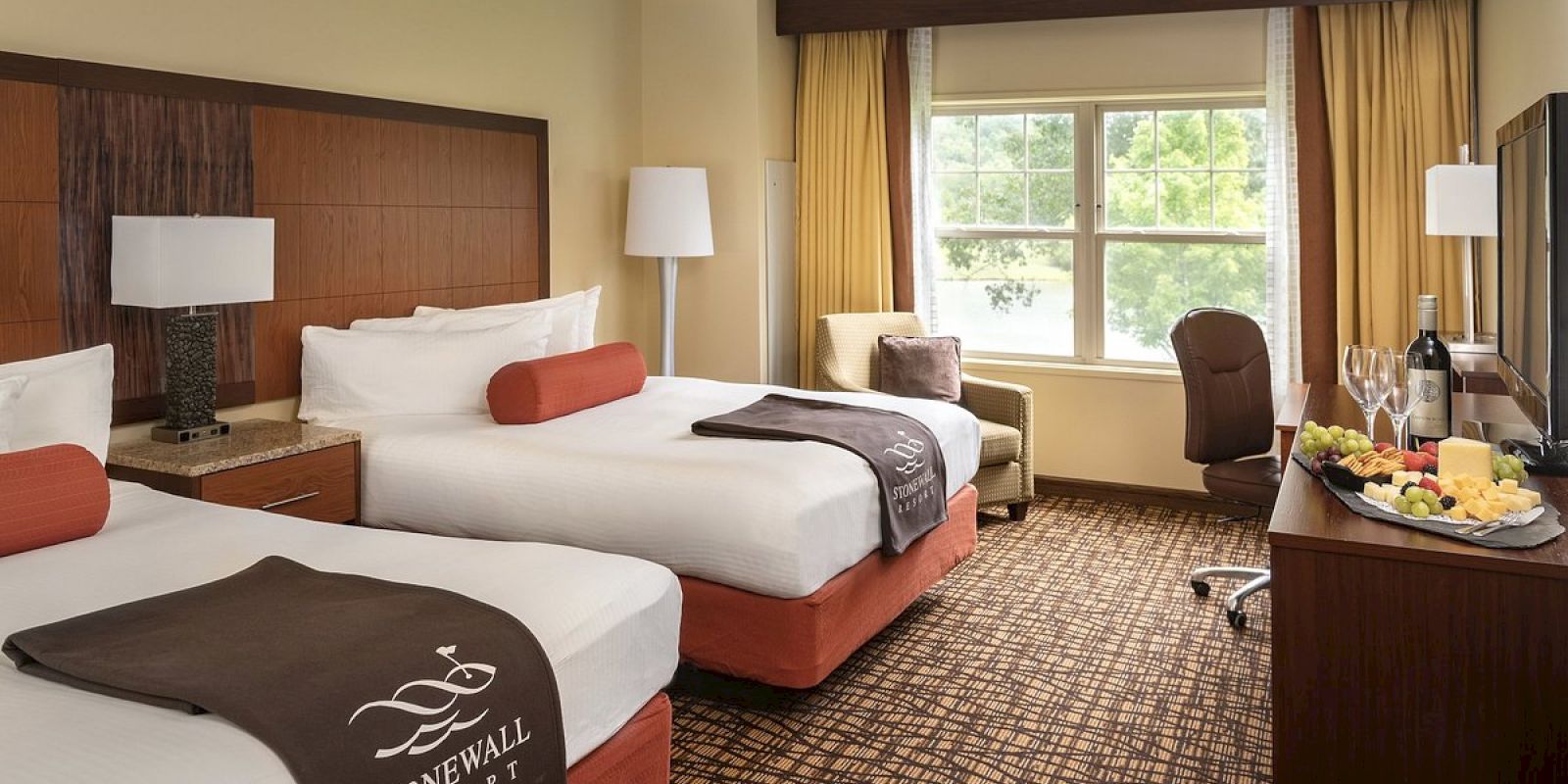 The image shows a hotel room with two double beds, a window with curtains, a chair, and a tray with wine and snacks on a TV stand.