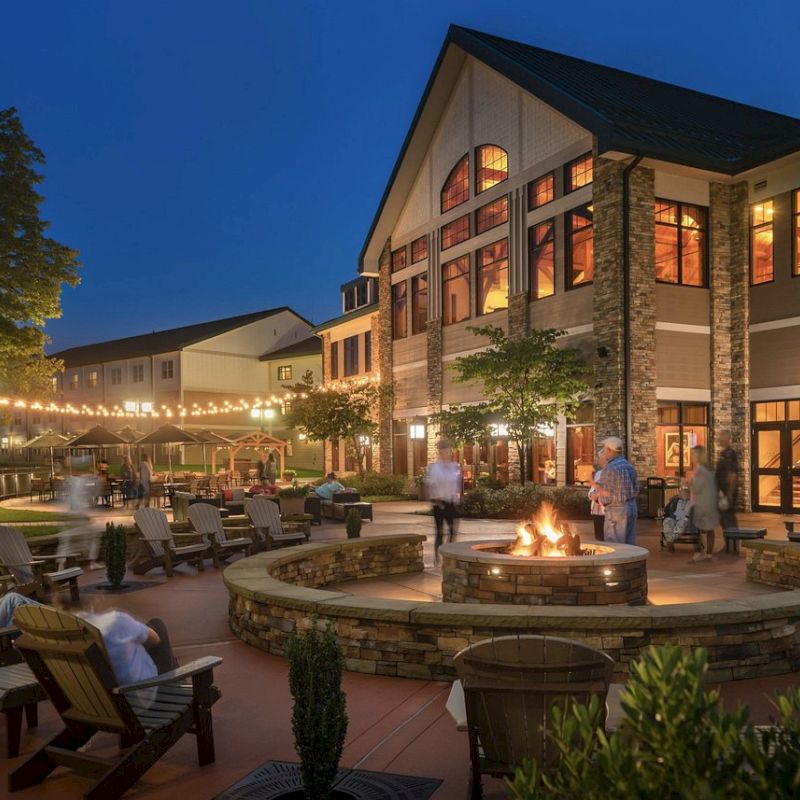 An outdoor gathering space with a fire pit, surrounded by chairs and people, in front of a well-lit building at night.