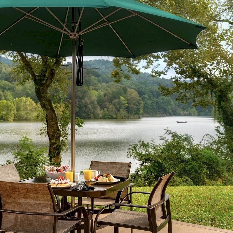 Outdoor dining area with a table set for breakfast under a green umbrella, overlooking a serene lake and lush greenery.