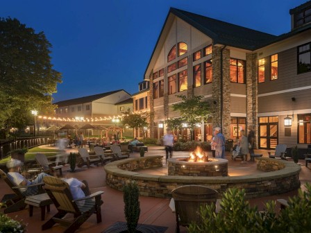 An outdoor gathering space with a fire pit, surrounded by chairs, people, and a building with lit windows and string lights in the evening.