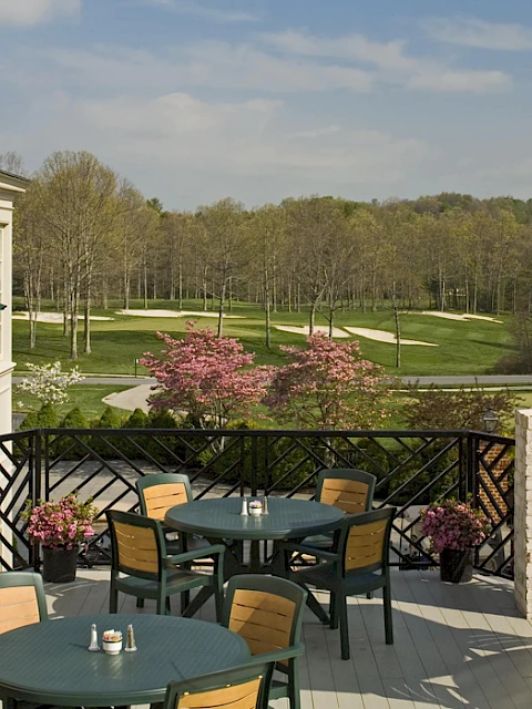 Outdoor patio with tables, chairs, umbrellas, and potted plants overlooking a golf course and blossoming trees.