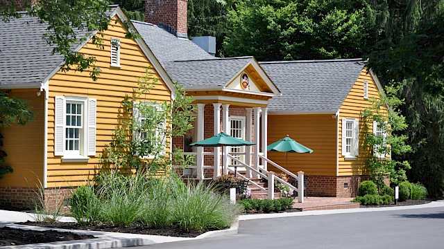 A charming yellow house with white shutters, a brick chimney, green umbrellas, and a lush garden surrounded by trees.