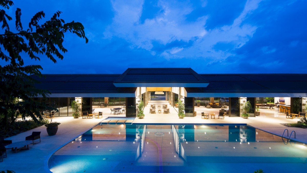 A luxurious building with an illuminated swimming pool, surrounded by seating areas and greenery, set against a twilight sky.