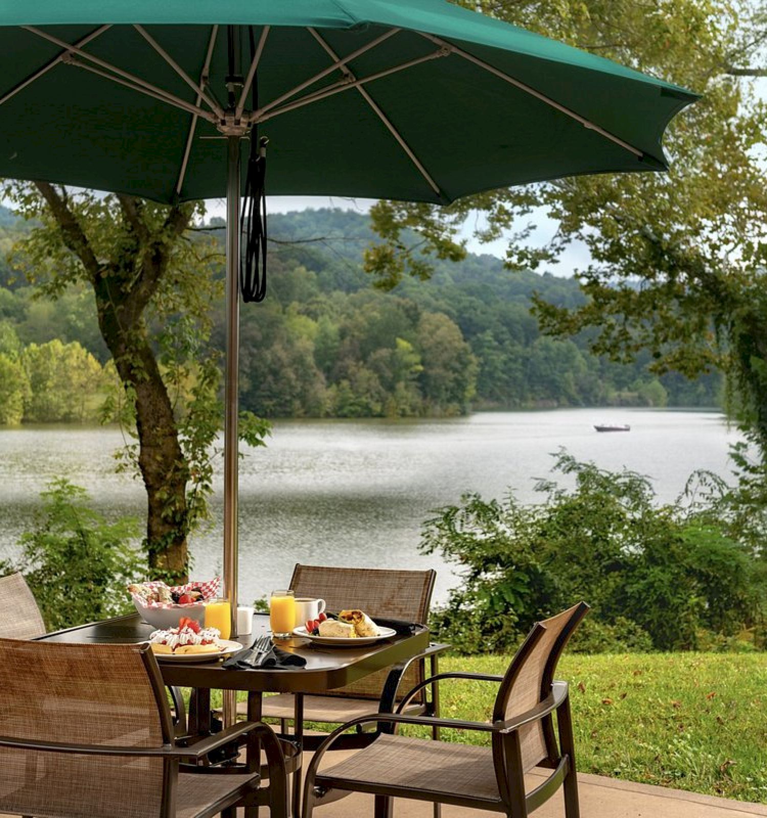 Outdoor dining setup with chairs and a table under a green umbrella, overlooking a scenic lake and lush greenery.