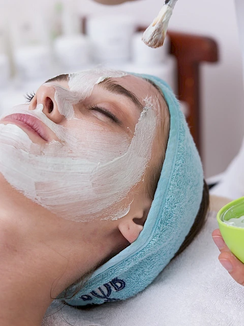 A woman is getting a facial treatment, with a beauty therapist applying a white cream or mask to her face.