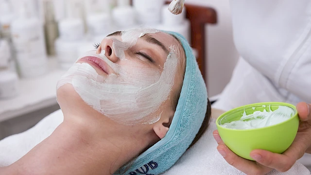 A woman is getting a facial treatment, with a beauty therapist applying a white cream or mask to her face.