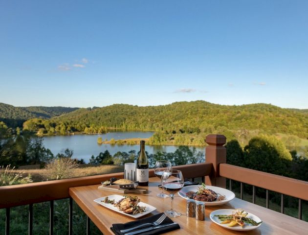A scenic view of a lake and hills from a balcony table set with wine, glasses, and plates of food in a peaceful outdoor dining setting.