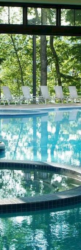 The image shows an indoor swimming pool area with large glass windows and sunlight streaming in. There are lounge chairs visible outside.